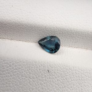 A 0.76-carat natural teal blue sapphire from Kanchanaburi, Thailand, with a unique pear-influenced oval faceted cut and saturated blue-green color, presented in a professional white jeweler's box.