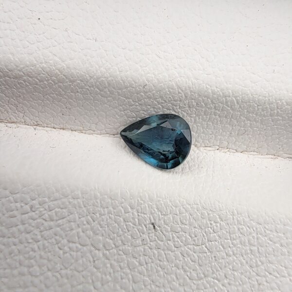 A 0.76-carat natural teal blue sapphire from Kanchanaburi, Thailand, with a unique pear-influenced oval faceted cut and saturated blue-green color, presented in a professional white jeweler's box.