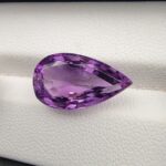 A premium 7.19-carat natural Brazilian amethyst (Katela) featuring a vibrant purple hue and a masterfully faceted pear shape, resting on a white textured jewelry display.