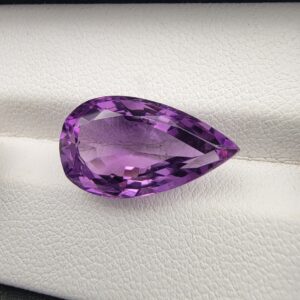A premium 7.19-carat natural Brazilian amethyst (Katela) featuring a vibrant purple hue and a masterfully faceted pear shape, resting on a white textured jewelry display.