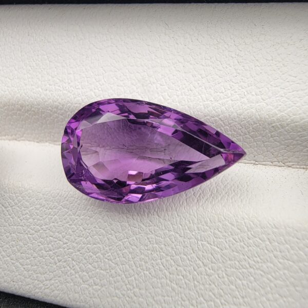 A premium 7.19-carat natural Brazilian amethyst (Katela) featuring a vibrant purple hue and a masterfully faceted pear shape, resting on a white textured jewelry display.