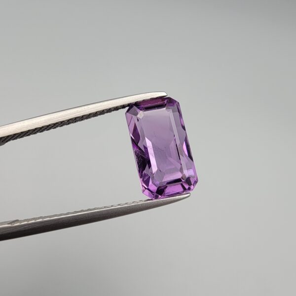 A natural 4.28-carat Brazilian amethyst (Katela) with a bright purple saturation and a sharp rectangle faceted cut, held in professional jeweler's tweezers.