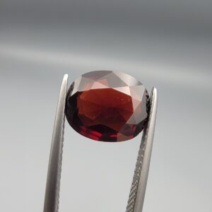 ​A natural 3.37-carat Red Garnet from Madagascar with a deep red sparkle and a polished oval faceted cut, held in professional jeweler's tweezers as shown in file 20260511_161141.jpg.