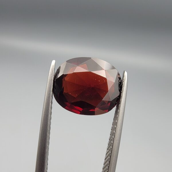 ​A natural 3.37-carat Red Garnet from Madagascar with a deep red sparkle and a polished oval faceted cut, held in professional jeweler's tweezers as shown in file 20260511_161141.jpg.