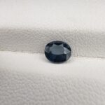 ​A natural 0.77-carat blue sapphire (Neelam) from Kanchanaburi, Thailand, featuring a deep navy blue hue and a masterfully faceted oval cut, resting on a white textured surface.