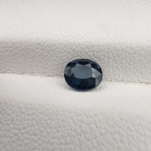 ​A natural 0.77-carat blue sapphire (Neelam) from Kanchanaburi, Thailand, featuring a deep navy blue hue and a masterfully faceted oval cut, resting on a white textured surface.