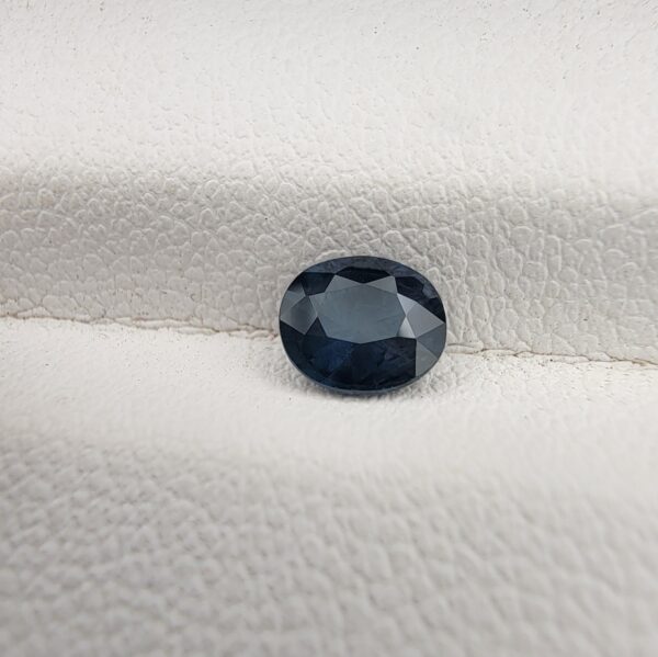 ​A natural 0.77-carat blue sapphire (Neelam) from Kanchanaburi, Thailand, featuring a deep navy blue hue and a masterfully faceted oval cut, resting on a white textured surface.