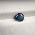 ​A natural 2.48-carat blue sapphire (Neelam) from Kanchanaburi, Thailand, featuring a deep navy blue hue and a sophisticated faceted pear shape, resting on a white textured background.