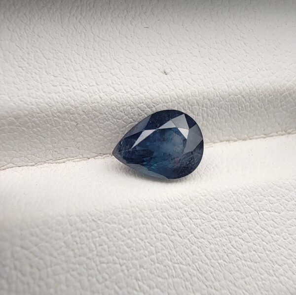 ​A natural 2.48-carat blue sapphire (Neelam) from Kanchanaburi, Thailand, featuring a deep navy blue hue and a sophisticated faceted pear shape, resting on a white textured background.