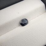 A natural 0.78-carat blue sapphire (Neelam) from Kanchanaburi, Thailand, featuring a deep navy blue hue and a masterfully faceted oval cut, resting on a white textured jewelry box.