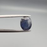 A natural 2.85-carat blue sapphire (Neelam) from Madagascar, featuring a deep navy blue hue and a masterfully faceted oval cut, held in professional jeweler's tweezers.
