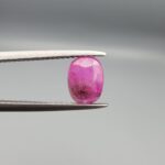 ​A natural 1.21-carat pinkish-red ruby from Mozambique featuring a faceted oval cut with a distinct dark natural inclusion, held in professional jeweler's tweezers.
