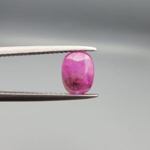 ​A natural 1.21-carat pinkish-red ruby from Mozambique featuring a faceted oval cut with a distinct dark natural inclusion, held in professional jeweler's tweezers.