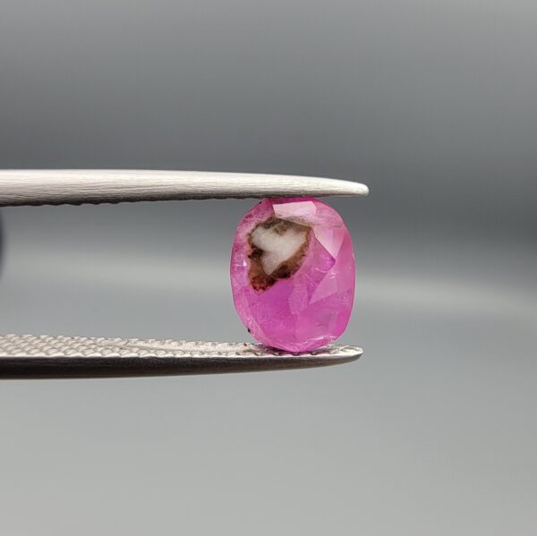 ​A natural 1.21-carat pinkish-red ruby from Mozambique featuring a faceted oval cut with a distinct dark natural inclusion, held in professional jeweler's tweezers.