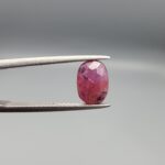 ​A natural 1.85-carat pinkish-red ruby from Mozambique featuring a faceted oval cut with visible natural internal silk and crystal inclusions, held in professional jeweler's tweezers.