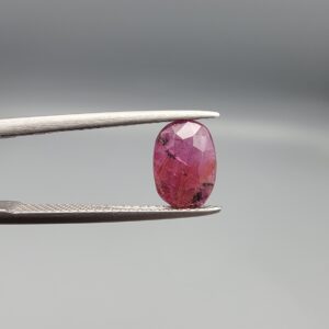 ​A natural 1.85-carat pinkish-red ruby from Mozambique featuring a faceted oval cut with visible natural internal silk and crystal inclusions, held in professional jeweler's tweezers.