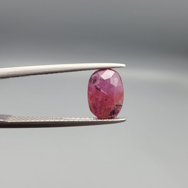 ​A natural 1.85-carat pinkish-red ruby from Mozambique featuring a faceted oval cut with visible natural internal silk and crystal inclusions, held in professional jeweler's tweezers.