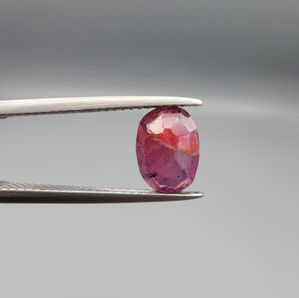 ​A natural 1.85-carat pinkish-red ruby from Mozambique featuring a faceted oval cut with visible natural internal silk and crystal inclusions, held in professional jeweler's tweezers.