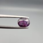 ​A natural 1.85-carat pinkish-red ruby from Mozambique featuring a faceted oval cut with visible natural internal silk and crystal inclusions, held in professional jeweler's tweezers.