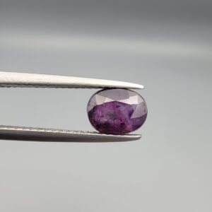 ​A natural 1.85-carat pinkish-red ruby from Mozambique featuring a faceted oval cut with visible natural internal silk and crystal inclusions, held in professional jeweler's tweezers.