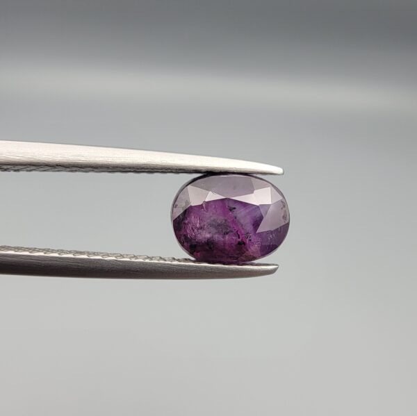 ​A natural 1.85-carat pinkish-red ruby from Mozambique featuring a faceted oval cut with visible natural internal silk and crystal inclusions, held in professional jeweler's tweezers.