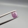 ​A natural 0.97-carat pinkish-red ruby from Mozambique featuring a faceted oval cut with visible natural internal birthmarks and silky inclusions, held in professional jeweler's tweezers.