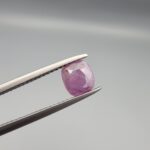 ​A natural 0.97-carat pinkish-red ruby from Mozambique featuring a faceted oval cut with visible natural internal birthmarks and silky inclusions, held in professional jeweler's tweezers.