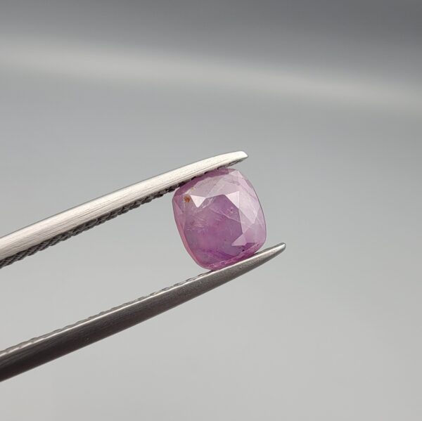 ​A natural 0.97-carat pinkish-red ruby from Mozambique featuring a faceted oval cut with visible natural internal birthmarks and silky inclusions, held in professional jeweler's tweezers.