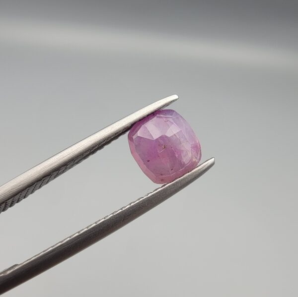 ​A natural 0.97-carat pinkish-red ruby from Mozambique featuring a faceted oval cut with visible natural internal birthmarks and silky inclusions, held in professional jeweler's tweezers.
