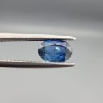 A natural 0.83-carat blue sapphire (Neelam) from Ceylon, Sri Lanka, featuring a faceted oval cut with a bright cornflower blue saturation, held in professional jeweler's tweezers.
