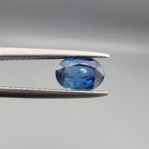 A natural 0.83-carat blue sapphire (Neelam) from Ceylon, Sri Lanka, featuring a faceted oval cut with a bright cornflower blue saturation, held in professional jeweler's tweezers.