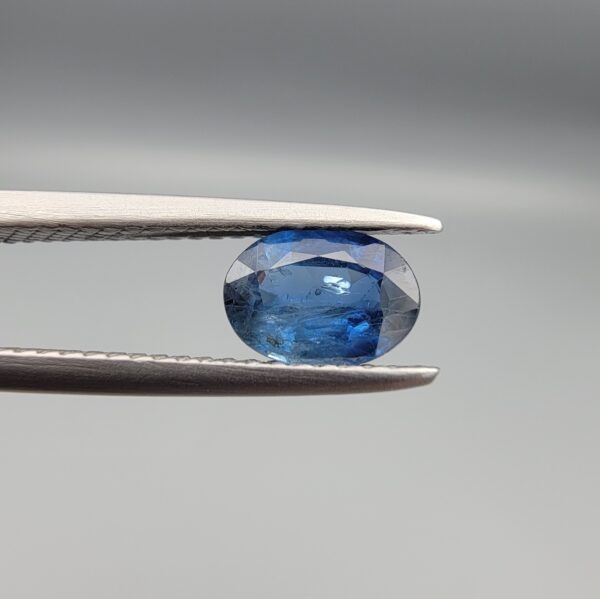 A natural 0.83-carat blue sapphire (Neelam) from Ceylon, Sri Lanka, featuring a faceted oval cut with a bright cornflower blue saturation, held in professional jeweler's tweezers.