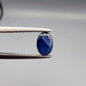 A natural 0.51-carat blue sapphire (Neelam) from Ceylon, Sri Lanka, featuring a faceted oval cut with bright cornflower blue color and high clarity, held in professional jeweler's tweezers.
