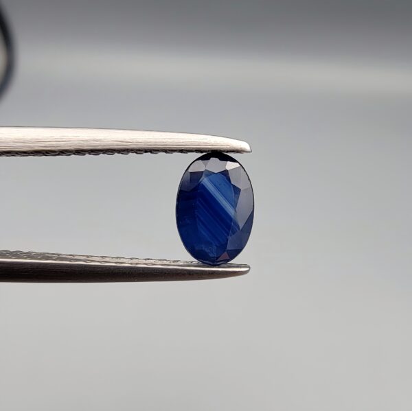 A natural 0.51-carat blue sapphire (Neelam) from Ceylon, Sri Lanka, featuring a faceted oval cut with bright cornflower blue color and high clarity, held in professional jeweler's tweezers.