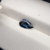 A natural 0.99-carat blue sapphire (Neelam) from Ceylon, Sri Lanka, featuring a masterfully faceted pear shape with brilliant cornflower blue color and a highly transparent crystal structure, resting elegantly on a textured white surface.