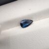 A natural 0.99-carat blue sapphire (Neelam) from Ceylon, Sri Lanka, featuring a masterfully faceted pear shape with brilliant cornflower blue color and a highly transparent crystal structure, resting elegantly on a textured white surface.