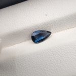 A natural 0.99-carat blue sapphire (Neelam) from Ceylon, Sri Lanka, featuring a masterfully faceted pear shape with brilliant cornflower blue color and a highly transparent crystal structure, resting elegantly on a textured white surface.
