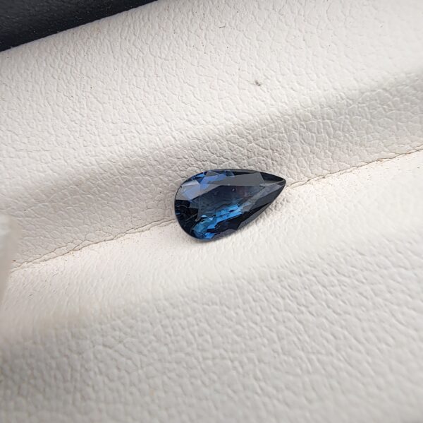 A natural 0.99-carat blue sapphire (Neelam) from Ceylon, Sri Lanka, featuring a masterfully faceted pear shape with brilliant cornflower blue color and a highly transparent crystal structure, resting elegantly on a textured white surface.