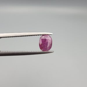 ​A natural 1.02-carat pinkish-red ruby (Yaqoot) from Mozambique, featuring a faceted oval cut with visible natural internal silk inclusions and character, held securely in professional jeweler's tweezers.
