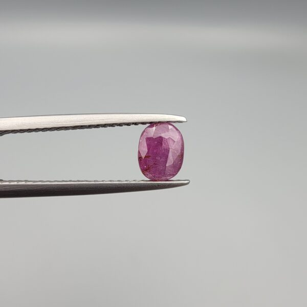 ​A natural 1.02-carat pinkish-red ruby (Yaqoot) from Mozambique, featuring a faceted oval cut with visible natural internal silk inclusions and character, held securely in professional jeweler's tweezers.