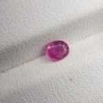 A natural 0.55-carat pinkish-red ruby (Yaqoot) from Mozambique, displaying a faceted oval cut with a distinct semi-transparent crystal structure and fine internal silk lines, resting on a textured white surface.