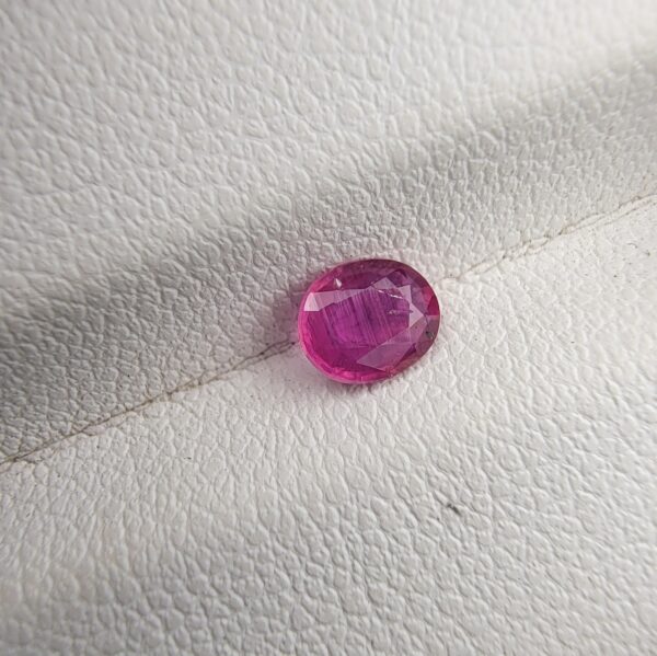 A natural 0.55-carat pinkish-red ruby (Yaqoot) from Mozambique, displaying a faceted oval cut with a distinct semi-transparent crystal structure and fine internal silk lines, resting on a textured white surface.