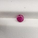 ​A natural 0.79-carat pinkish-red ruby (Yaqoot) from Mozambique, displaying a brilliant faceted round cut with a distinct translucent crystal structure and natural internal inclusions, resting on a textured white surface.