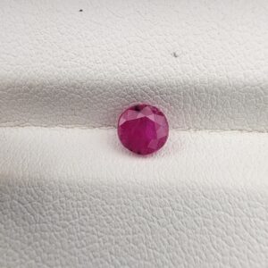​A natural 0.79-carat pinkish-red ruby (Yaqoot) from Mozambique, displaying a brilliant faceted round cut with a distinct translucent crystal structure and natural internal inclusions, resting on a textured white surface.