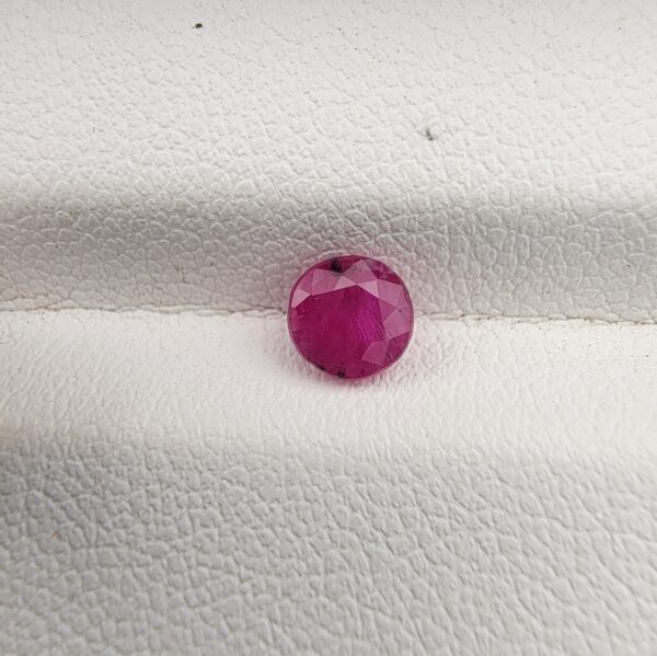 ​A natural 0.79-carat pinkish-red ruby (Yaqoot) from Mozambique, displaying a brilliant faceted round cut with a distinct translucent crystal structure and natural internal inclusions, resting on a textured white surface.