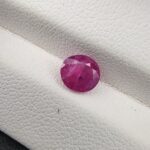 A natural 1.12-carat pinkish-red ruby (Yaqoot) from Mozambique, displaying a faceted oval cut with a distinct semi-transparent crystal structure and natural internal inclusions, resting on a textured white surface.