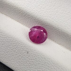 A natural 1.12-carat pinkish-red ruby (Yaqoot) from Mozambique, displaying a faceted oval cut with a distinct semi-transparent crystal structure and natural internal inclusions, resting on a textured white surface.