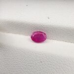 A natural 0.69-carat pinkish-red ruby (Yaqoot) from Mozambique, displaying a faceted oval cut with a distinct translucent crystal structure and natural internal inclusions, resting on a textured white surface.