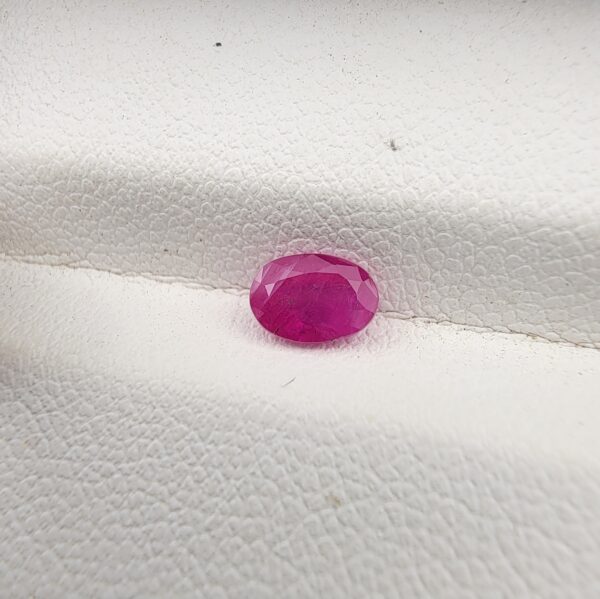 A natural 0.69-carat pinkish-red ruby (Yaqoot) from Mozambique, displaying a faceted oval cut with a distinct translucent crystal structure and natural internal inclusions, resting on a textured white surface.