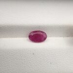 A natural 1.37-carat red ruby (Yaqoot) from Mozambique, displaying a faceted oval cut with a distinct translucent crystal structure and natural internal inclusions, resting on a textured white surface.